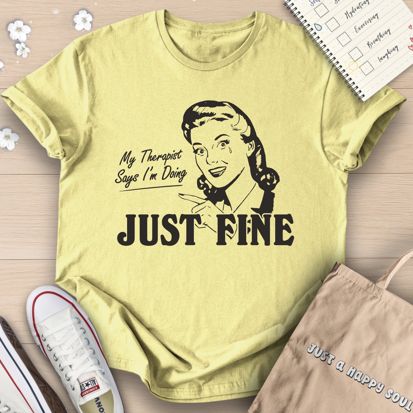 Yellow t-shirt with graphic and text 'My Therapist Says I'm Doing Just Fine' on a wooden surface.